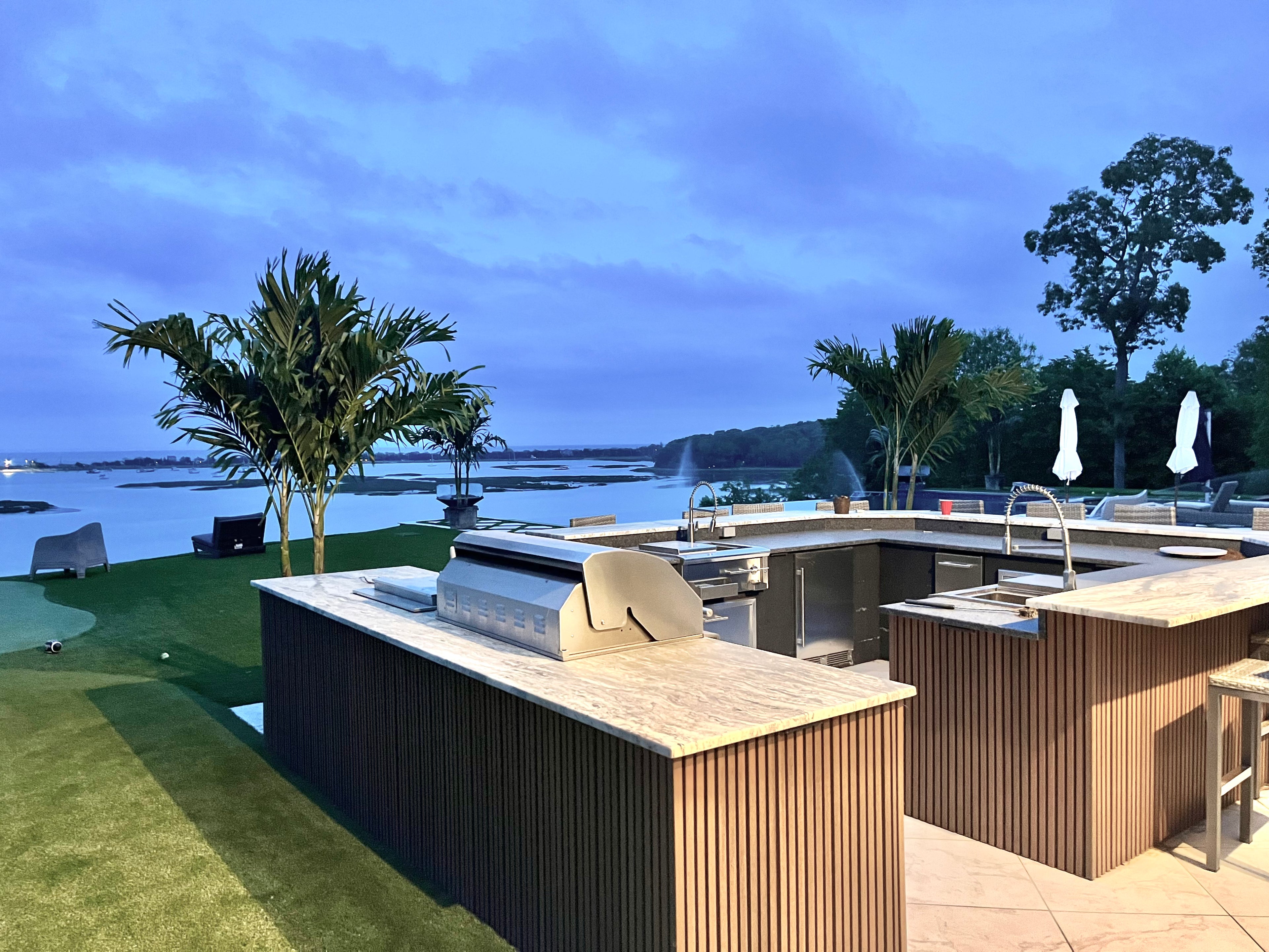 Outdoor kitchen area with grill and bar by a waterfront, featuring palm trees and a clear sky.